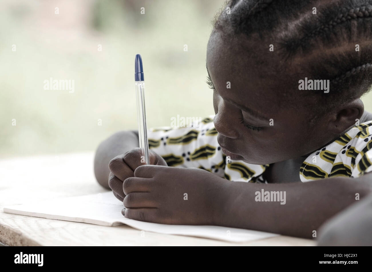 Cute African schoolgirl doing her homework at school: beautiful black girl writing and learning activity with a blue pen. She's sitting in her desk do Stock Photo