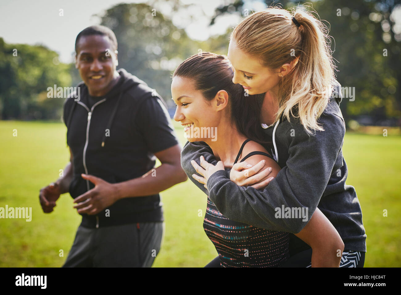 Personal trainer encouraging two women piggy backing in park Stock Photo