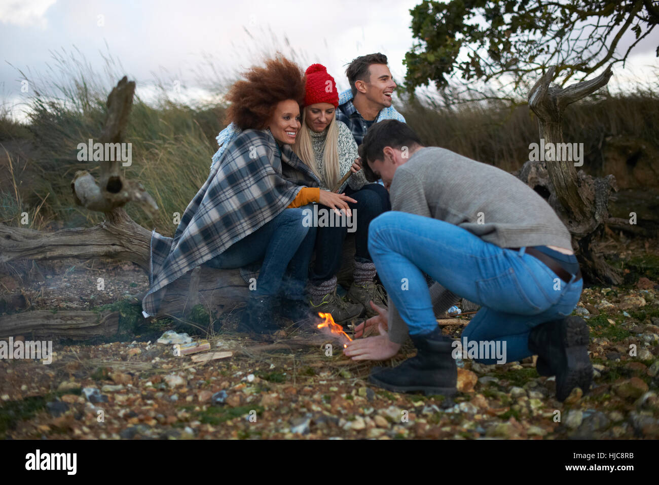 Four adult friends igniting campfire on windy beach Stock Photo