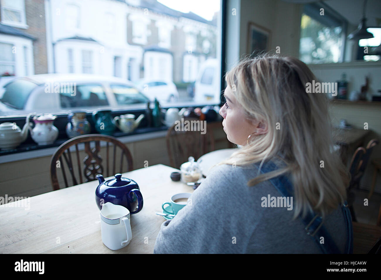 Young woman sitting in cafe, cup of tea and teapot on table, looking out of window Stock Photo