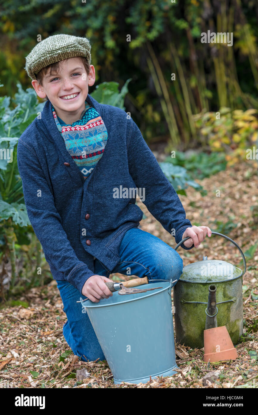 Happy smiling male boy child wearing hat or flat cap, gardening with bucket, garden fork and watering can in a vegetable patch Stock Photo