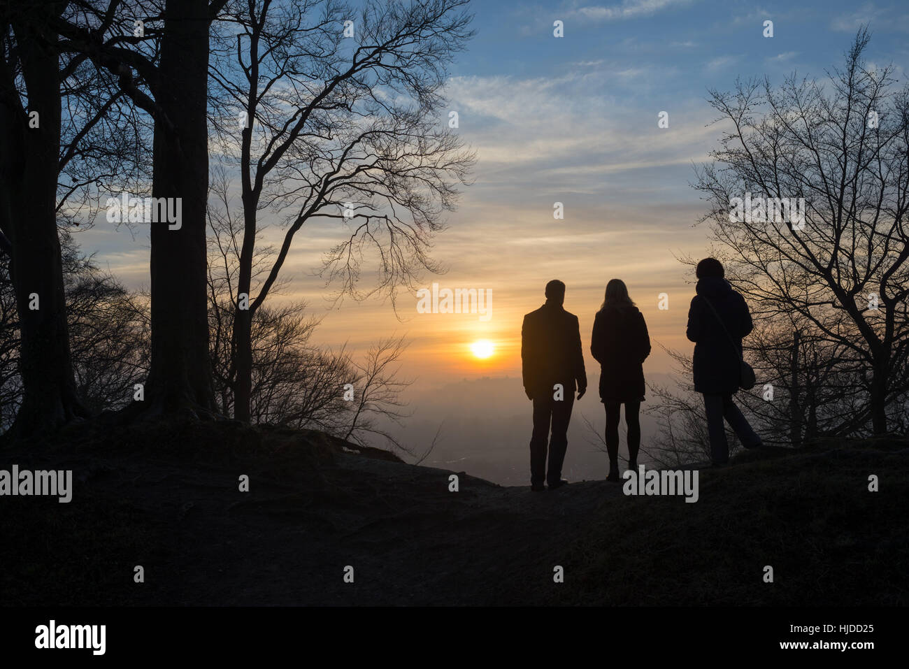 Group of people standing among trees looking at a misty winter sunset from high ground and silhouetted by the sun Stock Photo
