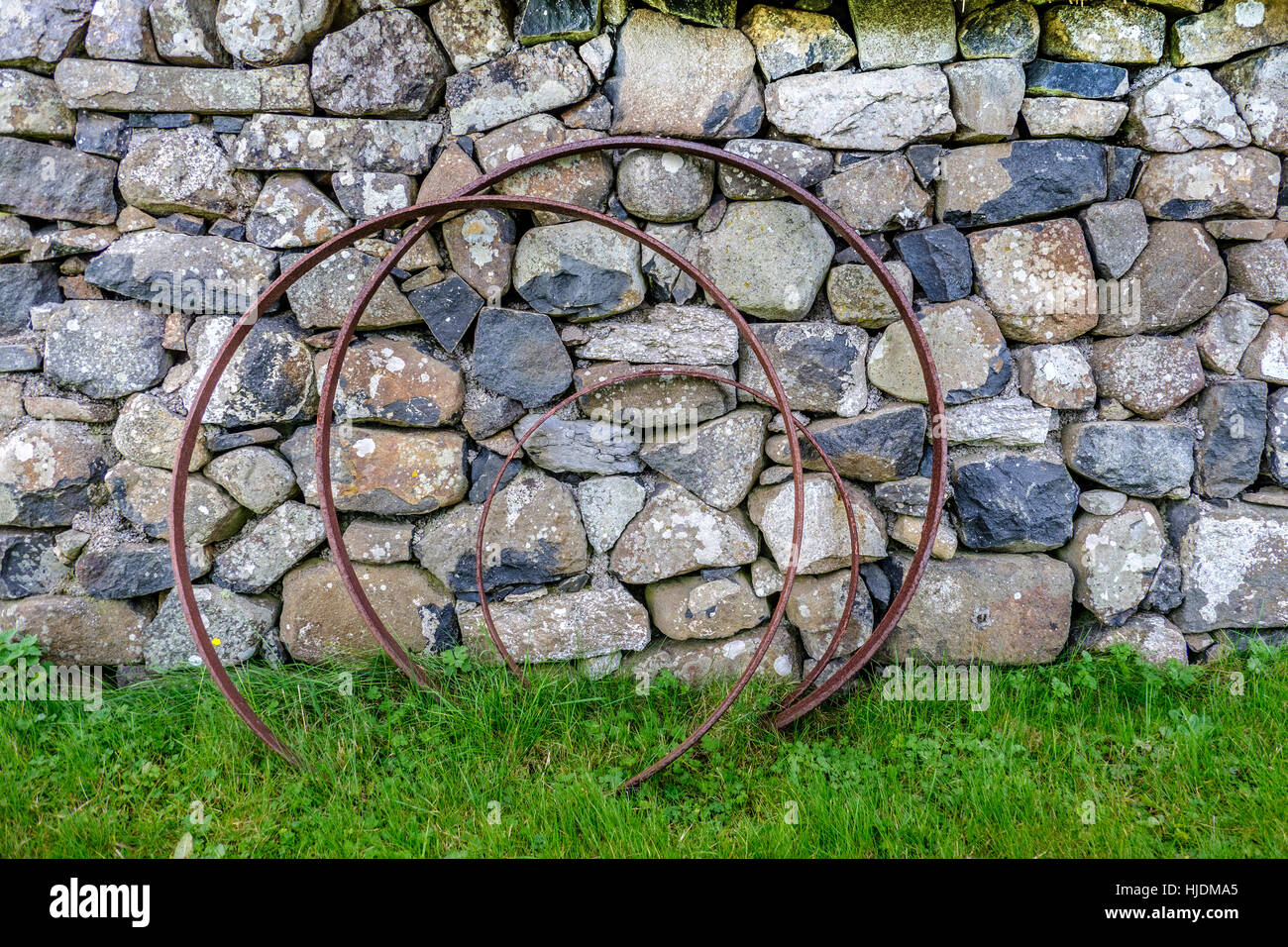 Rusting metal hoops against old lichen covered stone wall with grass and weeds foreground Stock Photo