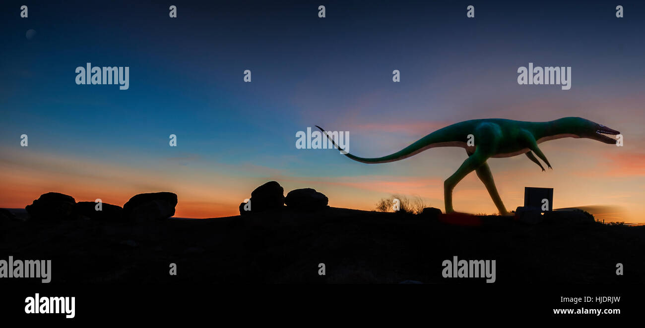 A concrete dinosaur overlooks Old Route 66 as a prismatic afterglow silhouettes the desert landscape.Panoramic image. Stock Photo