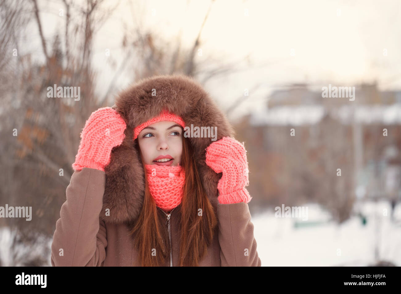 Lovely teen girl in pink knitted gloves, hat and scarf, fur hooded jacket winter walks in the park. She straightens the hood. Outdoor Activities. Stock Photo