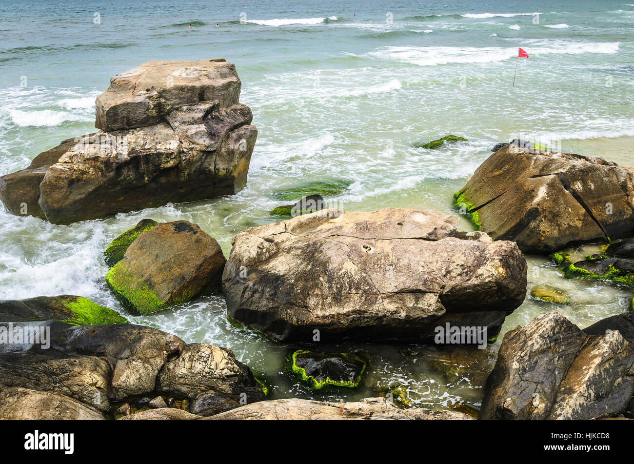 Beautiful scene with stones with green moss on the seashore, sea water passing between them, some small waves and people swimming in the background. Stock Photo