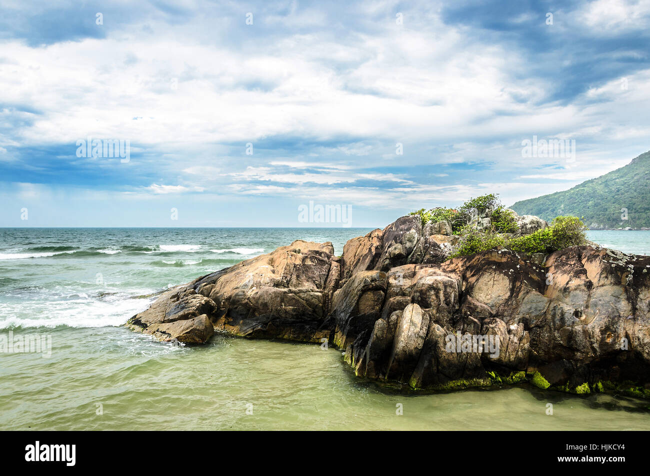 Rock formation in the middle of the sea. Waves hitting the rocks and a green mountain on the background. Stock Photo