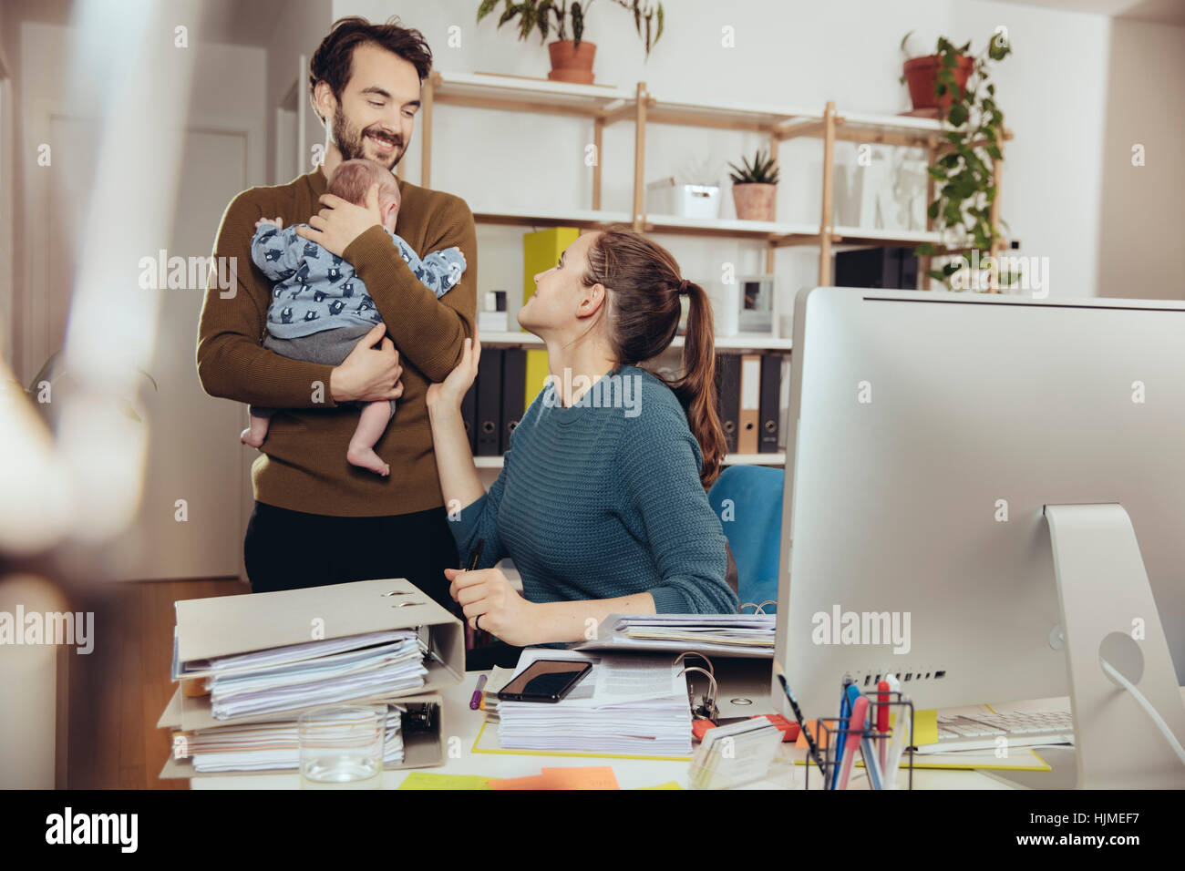 Smiling mother at desk looking at father holding baby in home office Stock Photo