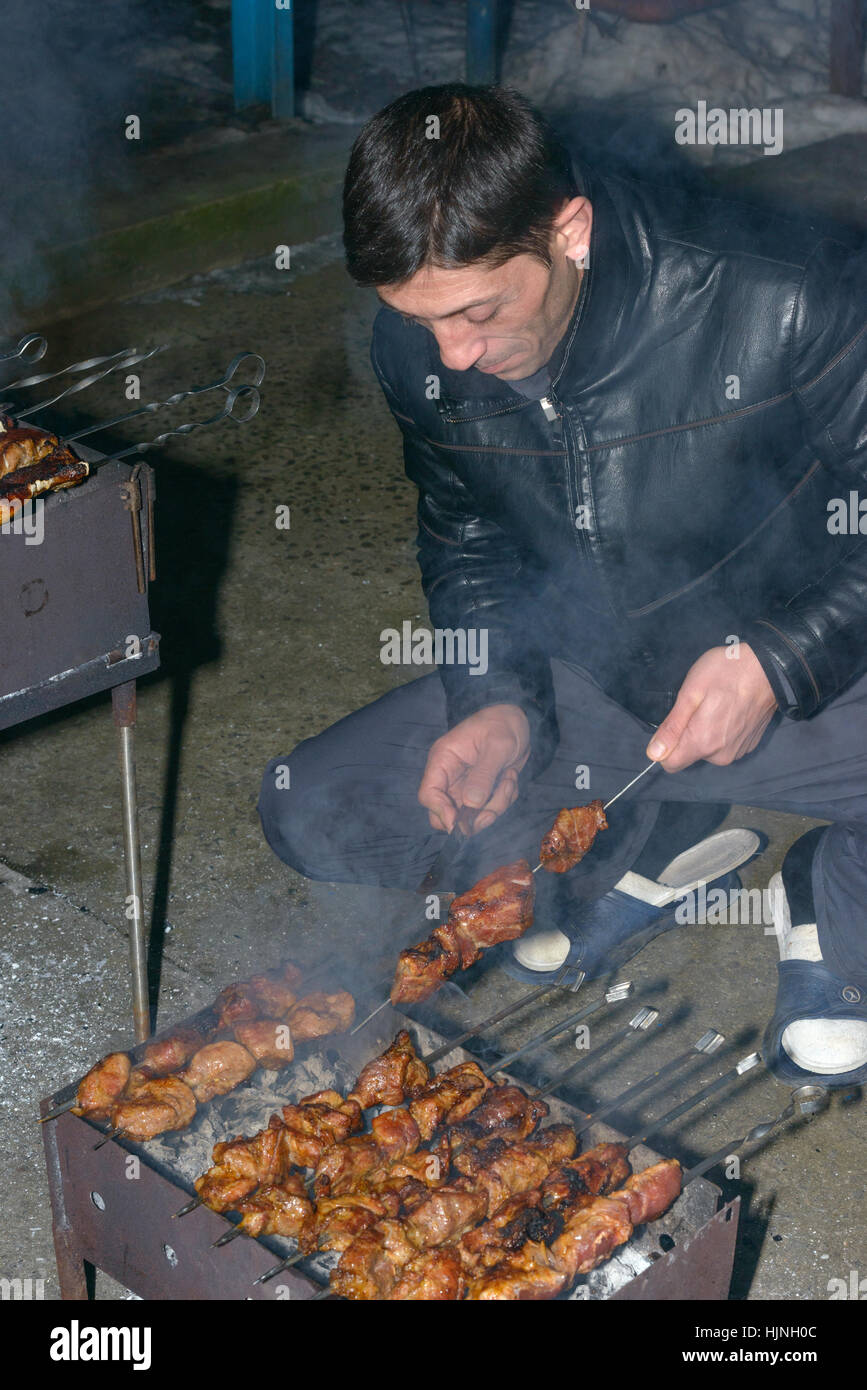 Mature man is cooking barbecued meat on compact metal mangal with skewers in evening. Stock Photo