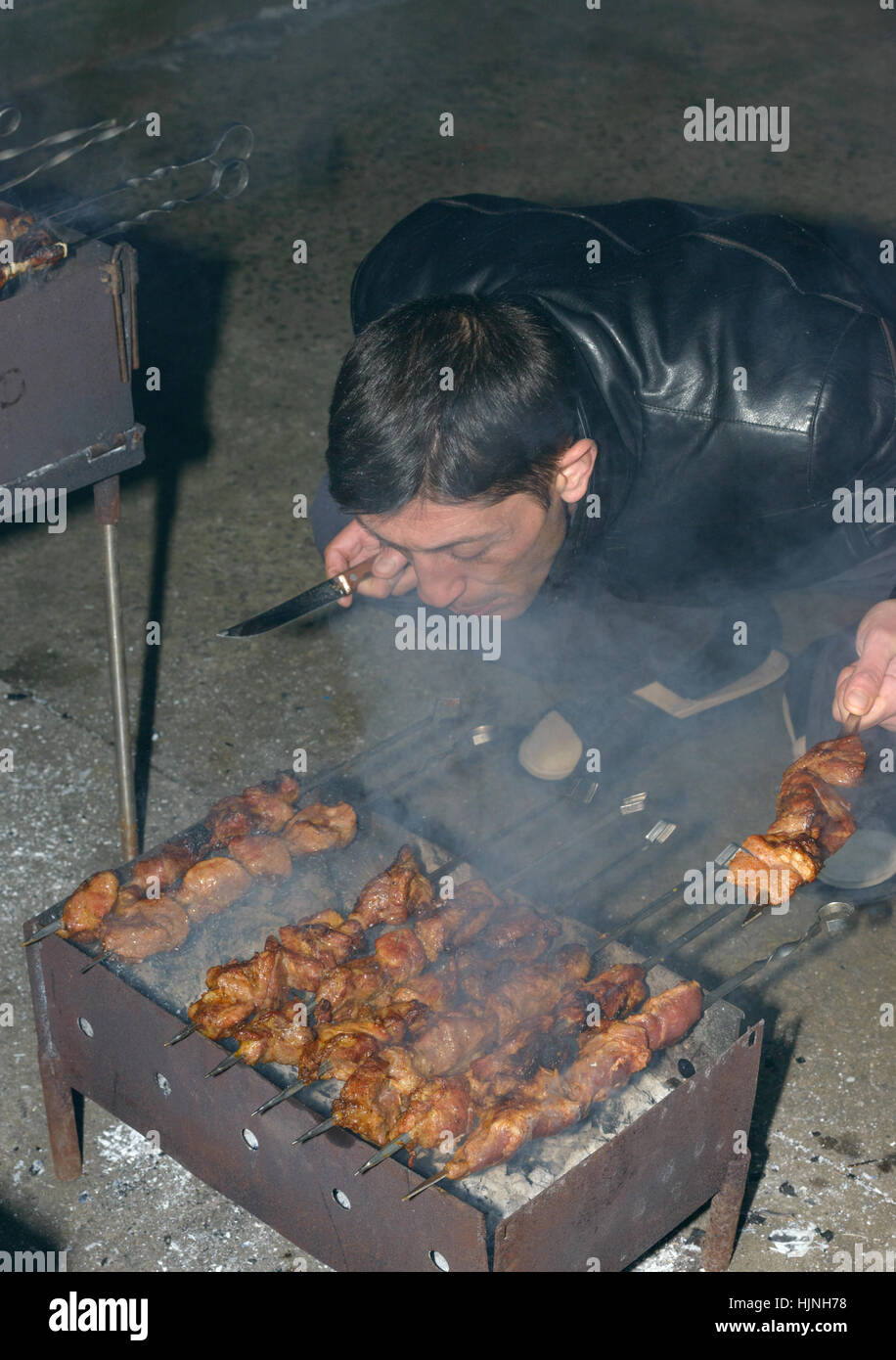 Mature man is cooking barbecued meat on compact metal mangal with skewers in evening. Stock Photo