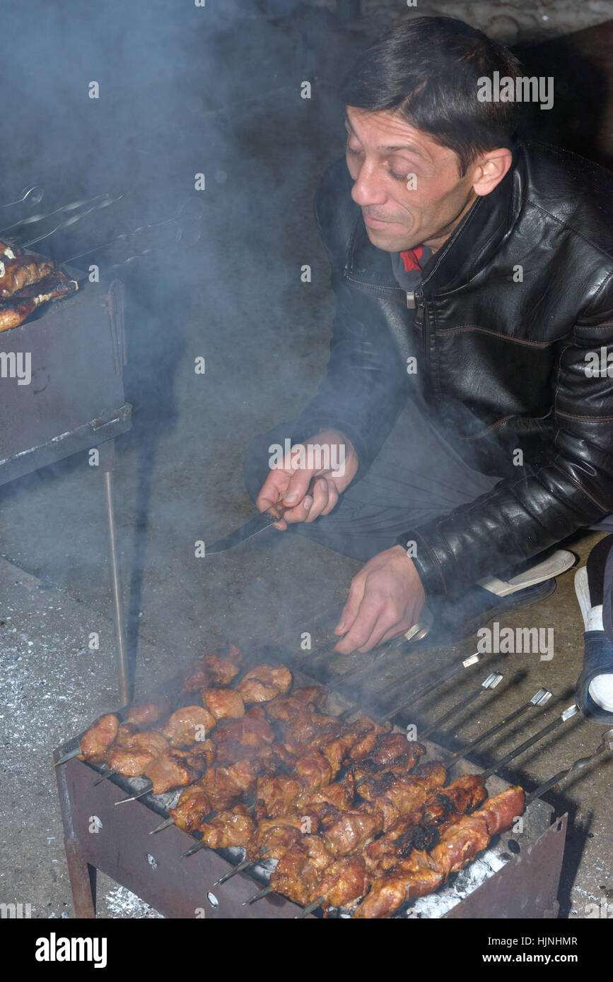 Mature man is cooking barbecued meat on compact metal mangal with skewers in evening. Stock Photo