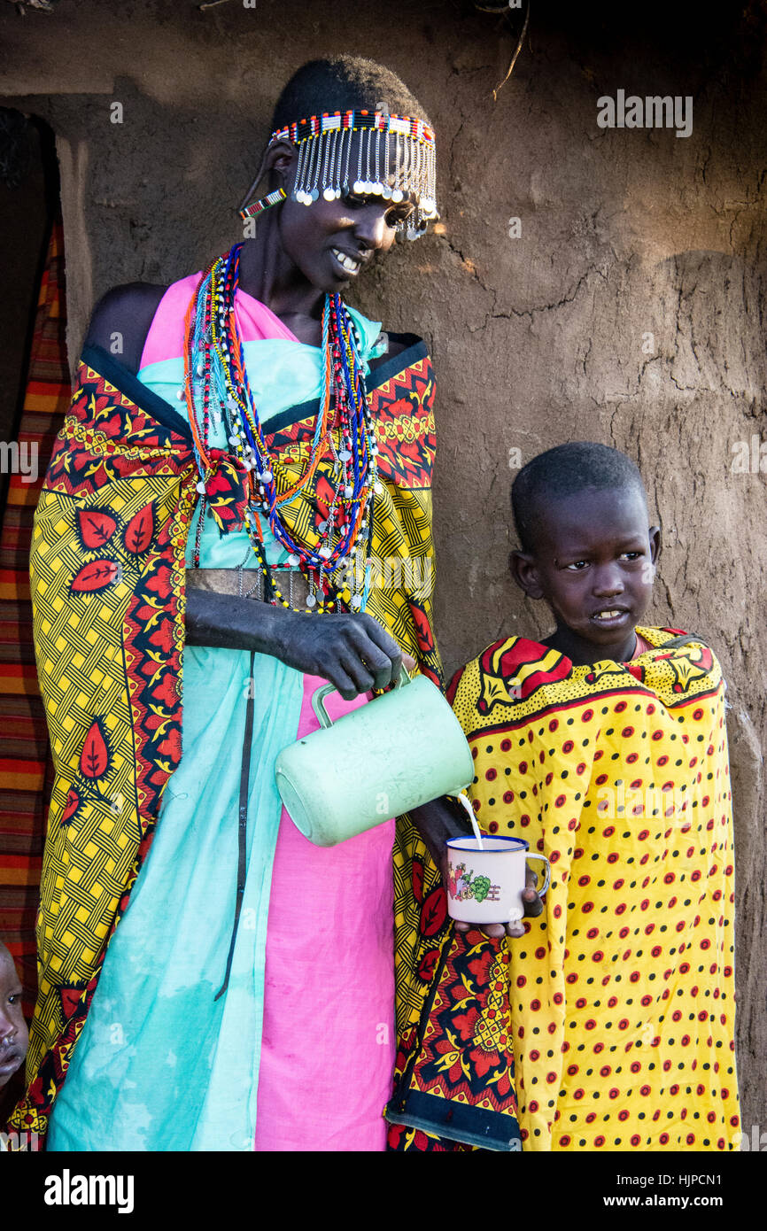 Maasai Woman pouring milk for a child, wearing traditional attire, in a village near the Masai Mara National Park, Kenya, East Africa Stock Photo