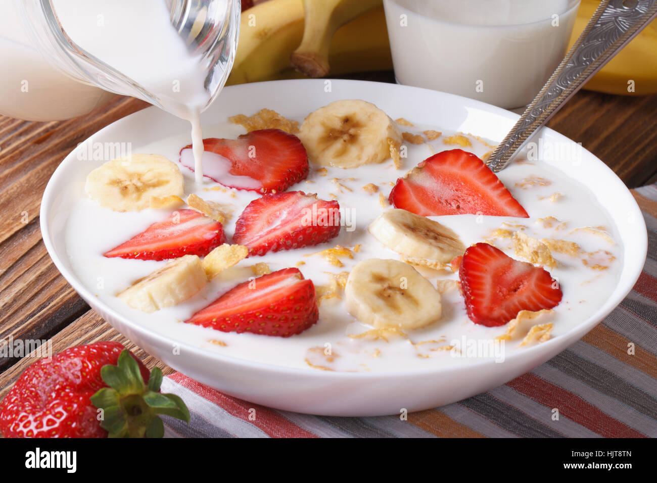 muesli with fresh strawberries and banana and milk is poured from a jug on the table closeup. horizontal Stock Photo