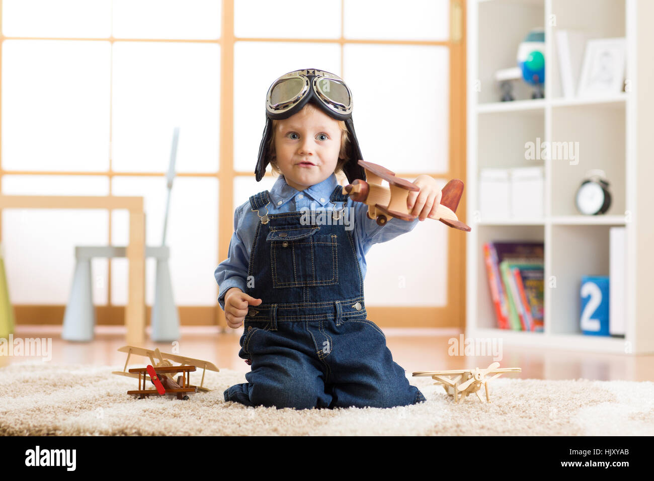 Kid boy weared aviator helmet plays with wooden toy plane in his children room Stock Photo