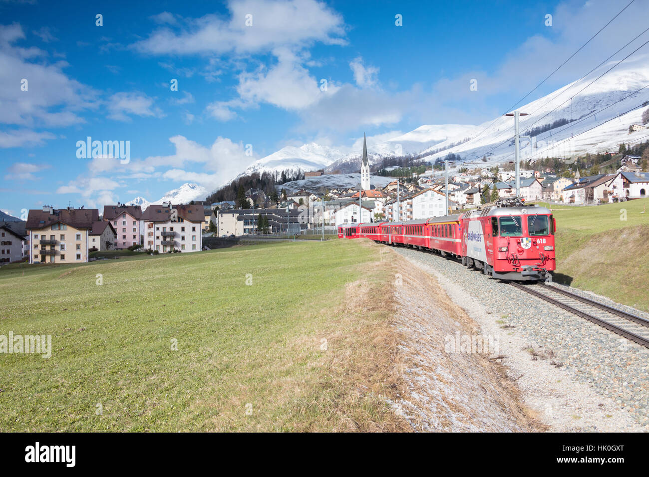 The red train runs across the alpine village of Zuoz in spring, Maloja, Canton of Graubunden, Engadine, Switzerland Stock Photo