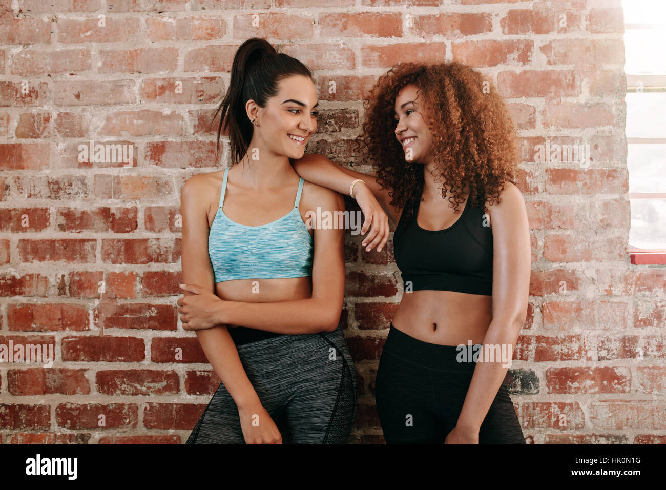 Portrait of two fitness females in gym relaxing and talking after workout. Young woman friends standing against wall and smiling. Stock Photo
