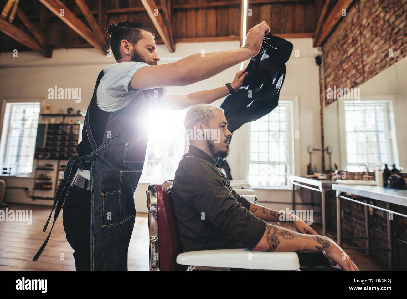 Hairstylist putting salon cape to customer before haircut. Male hairdresser at work with handsome man sitting on chair. Stock Photo