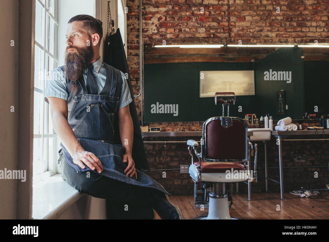 Young barber at his barbershop. Caucasian male with long beard sitting on sill of a window and looking outside. Stock Photo