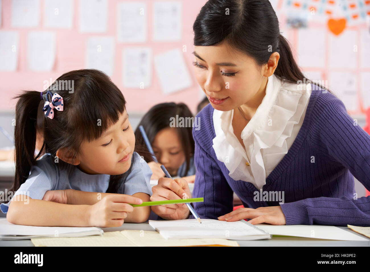 Teacher Helping Student Working At Desk In Chinese School Classroom ...
