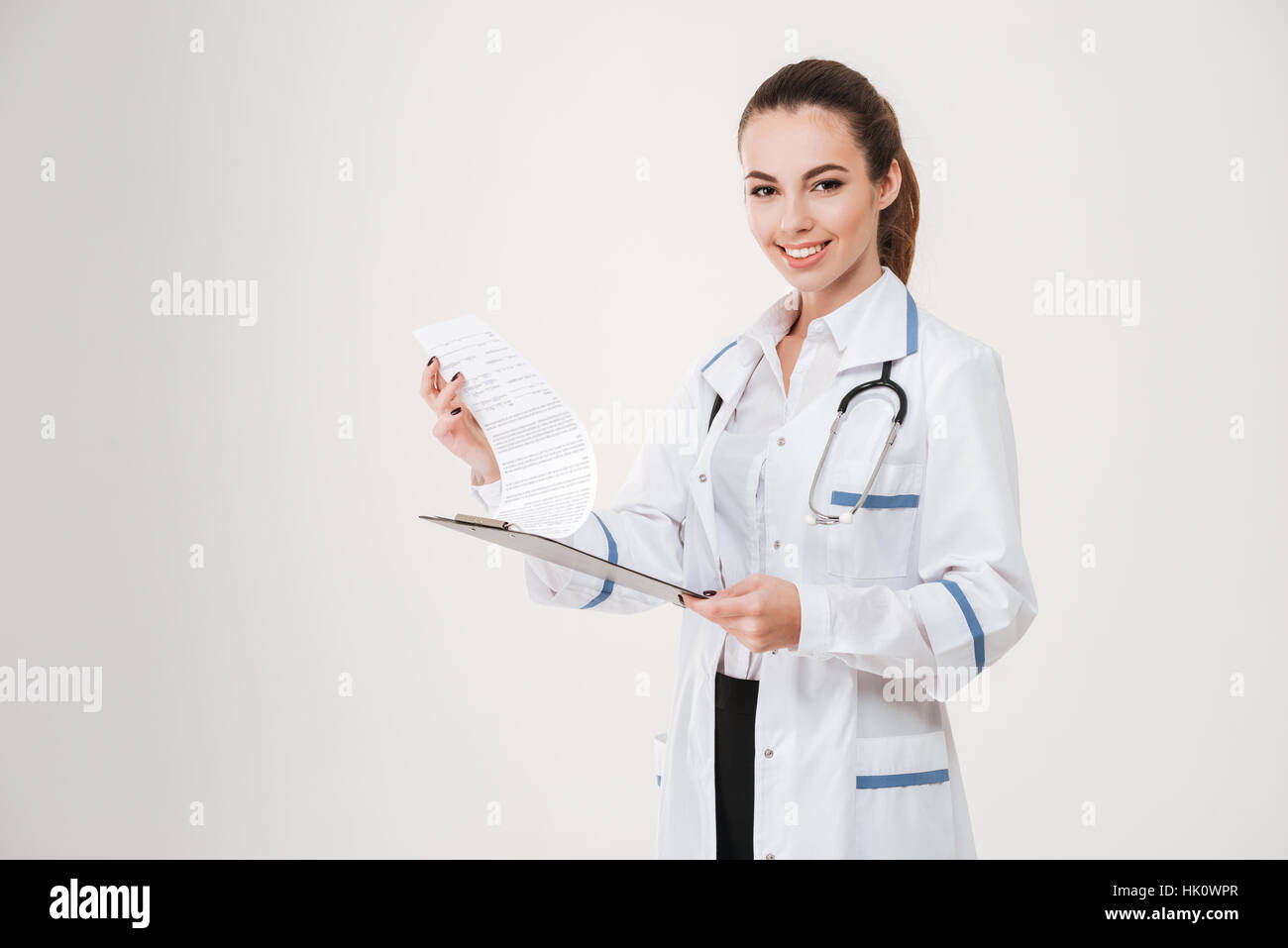 Cheerful pretty young woman doctor holding clipboard with documents over white background Stock Photo