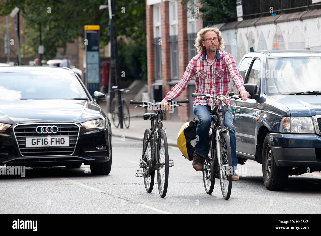 LONDON, ENGLAND - JULY 12, 2016 Serious blond curly bearded man in a plaid shirt riding a bike and holding a second bike for the wheel in Shoreditch. Stock Photo