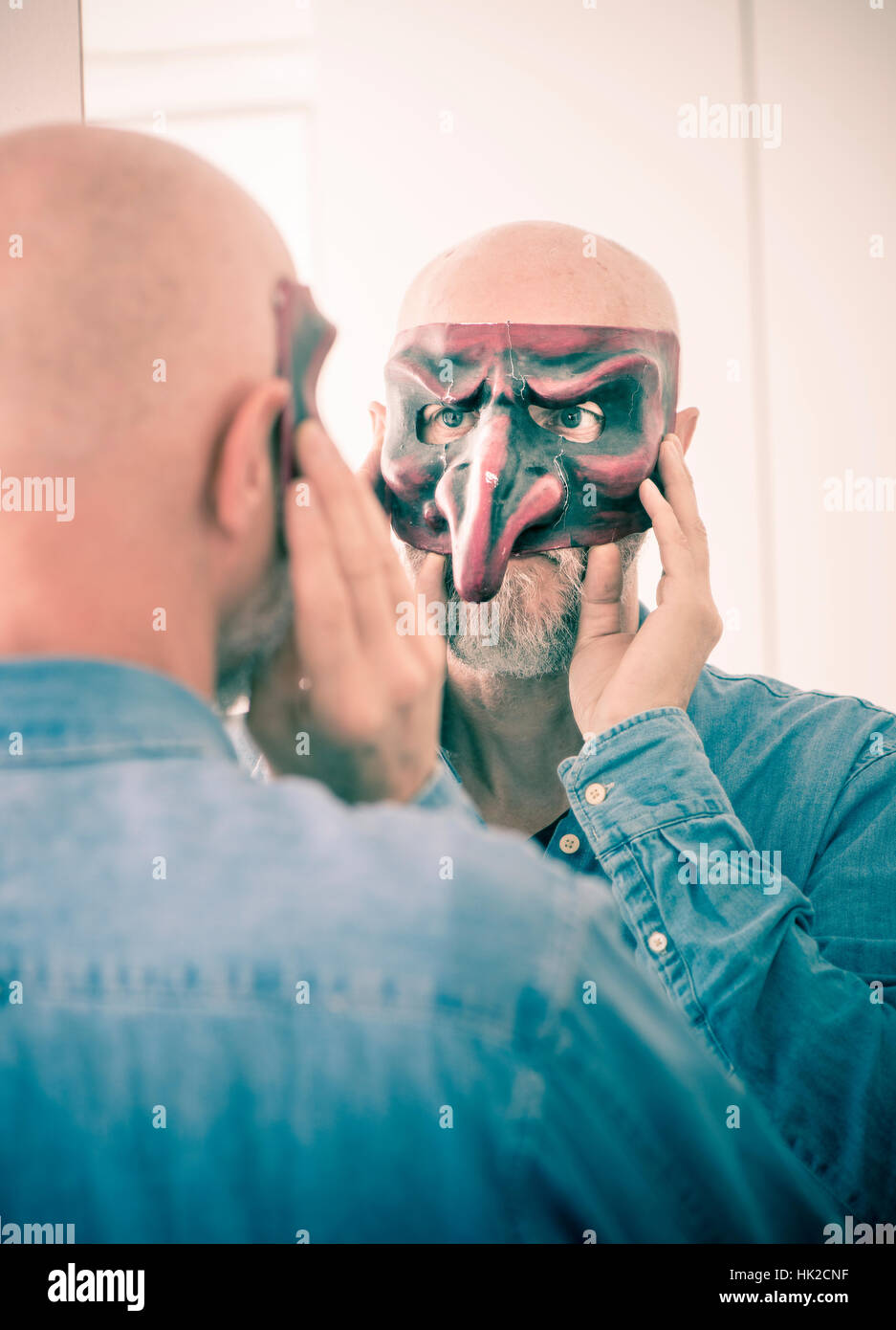 Old man cover face with carnival mask. Looking at himself in a mirror. Concept of ageing, self image as a senior person and hiding identity. Stock Photo