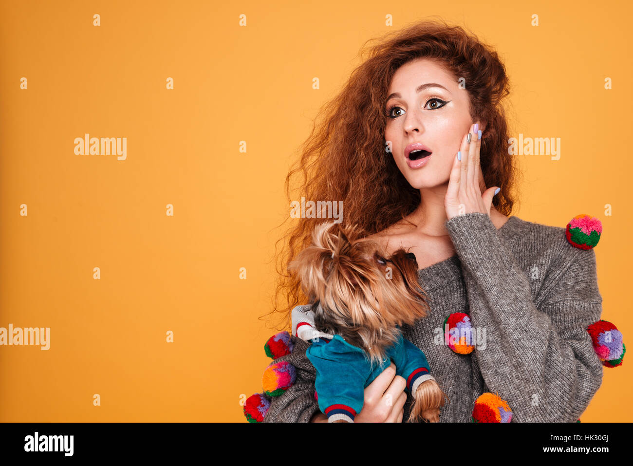 Young red head woman looking surprised while holding her dog isolated on orange background Stock Photo