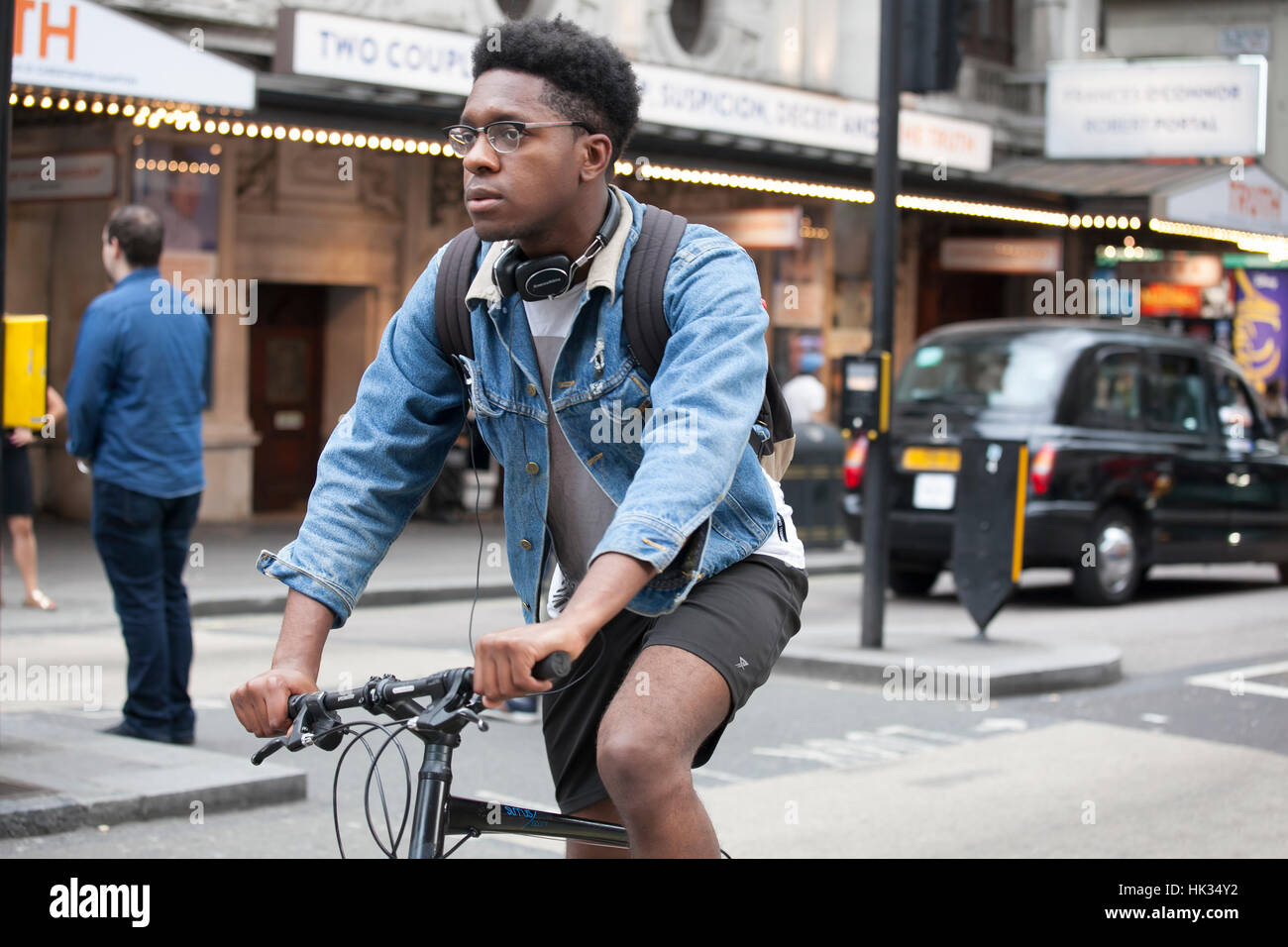 LONDON, ENGLAND - JULY 12, 2016 Serious young man in a denim jacket and short shorts riding a bicycle on the street Stock Photo