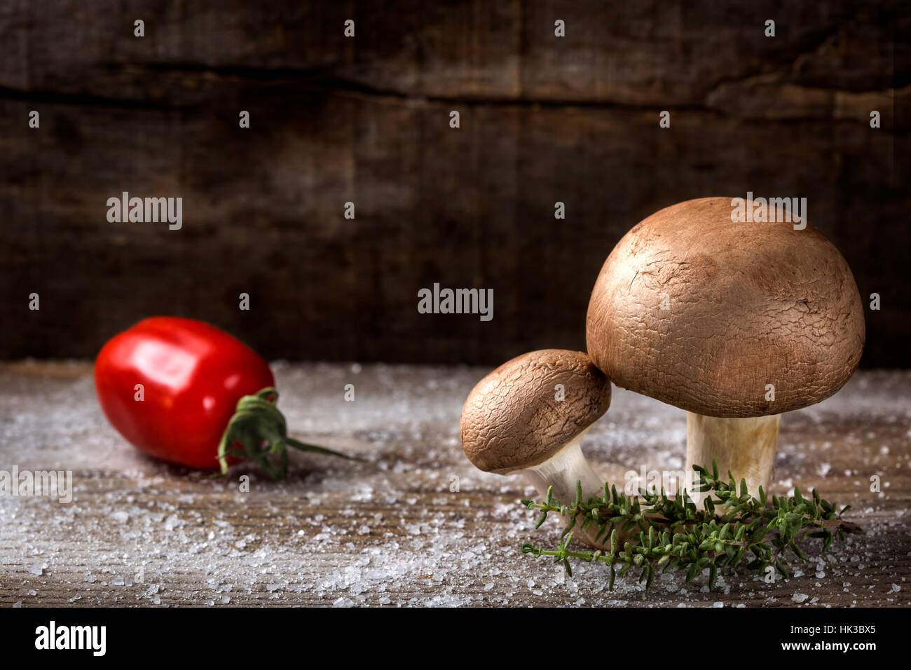 Two brown mushrooms with salt, thyme and one tomato out of focus over wooden background Stock Photo