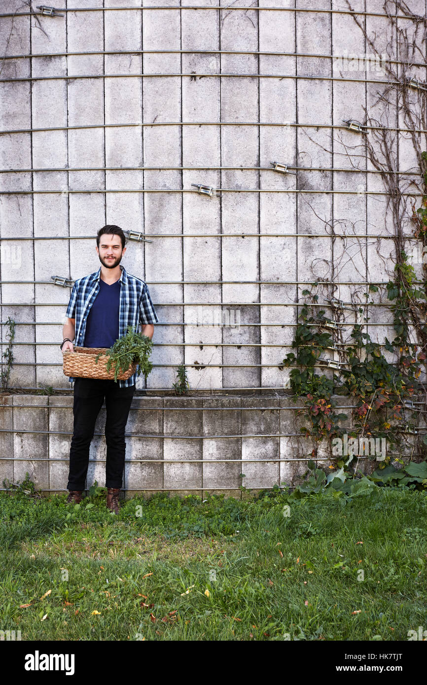 A young man in working clothes standing in front of a wall with plants growing up a trellis, holding a basket of crops. Stock Photo