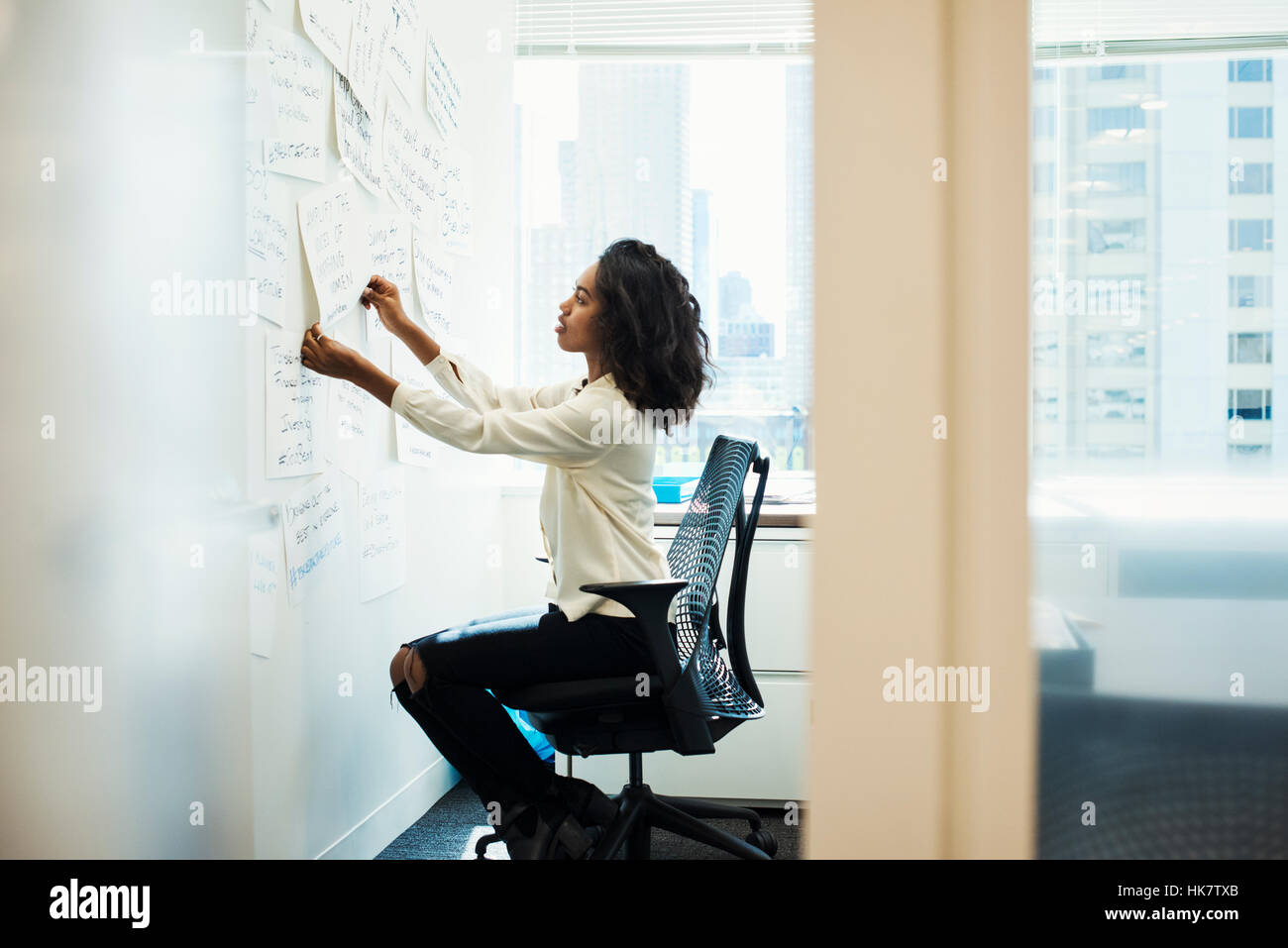 A woman sitting on a chair in an office arranging pieces of paper pinned on a whiteboard. Stock Photo