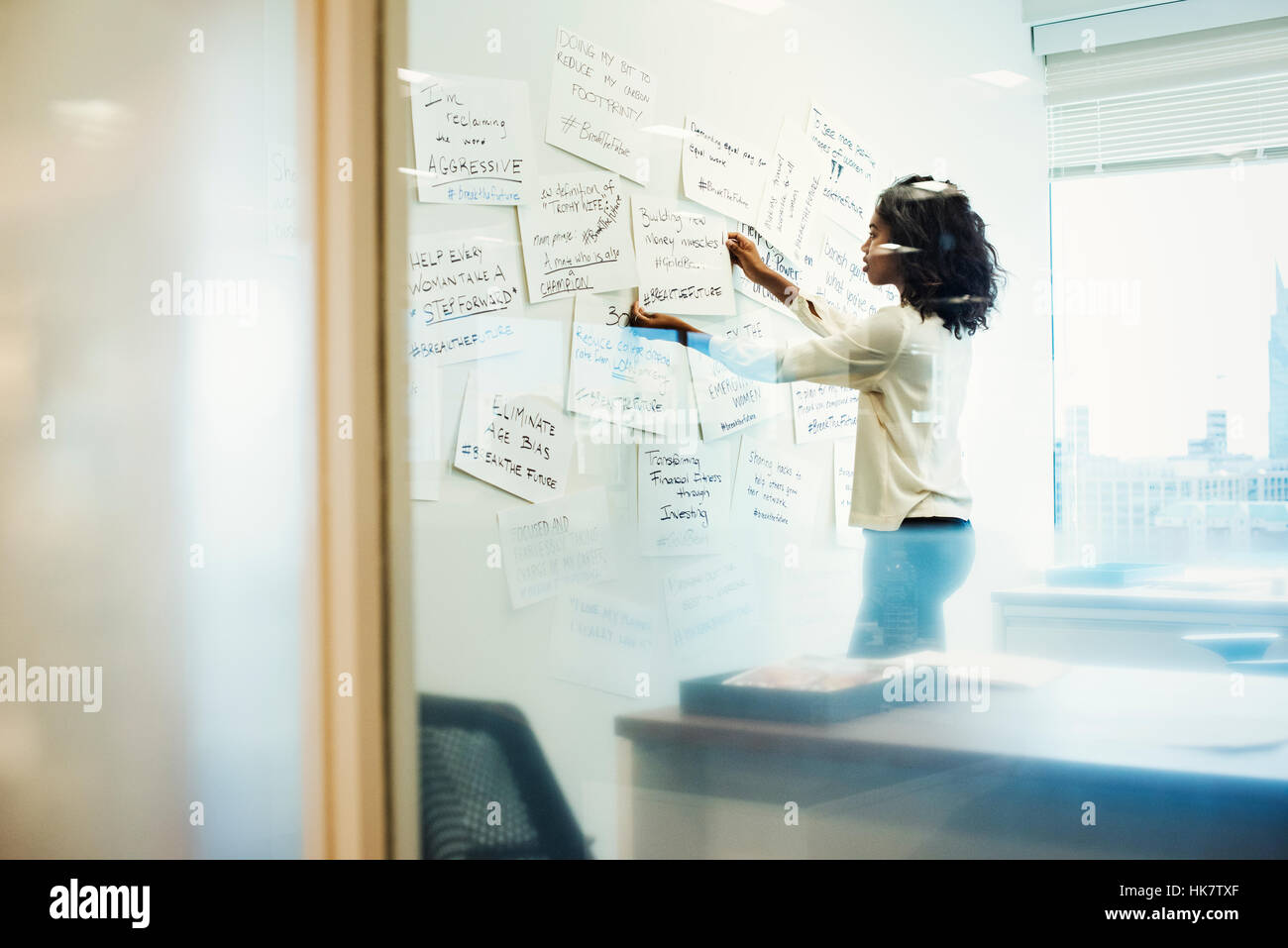 A woman standing in an office arranging pieces of paper pinned on a whiteboard. Stock Photo