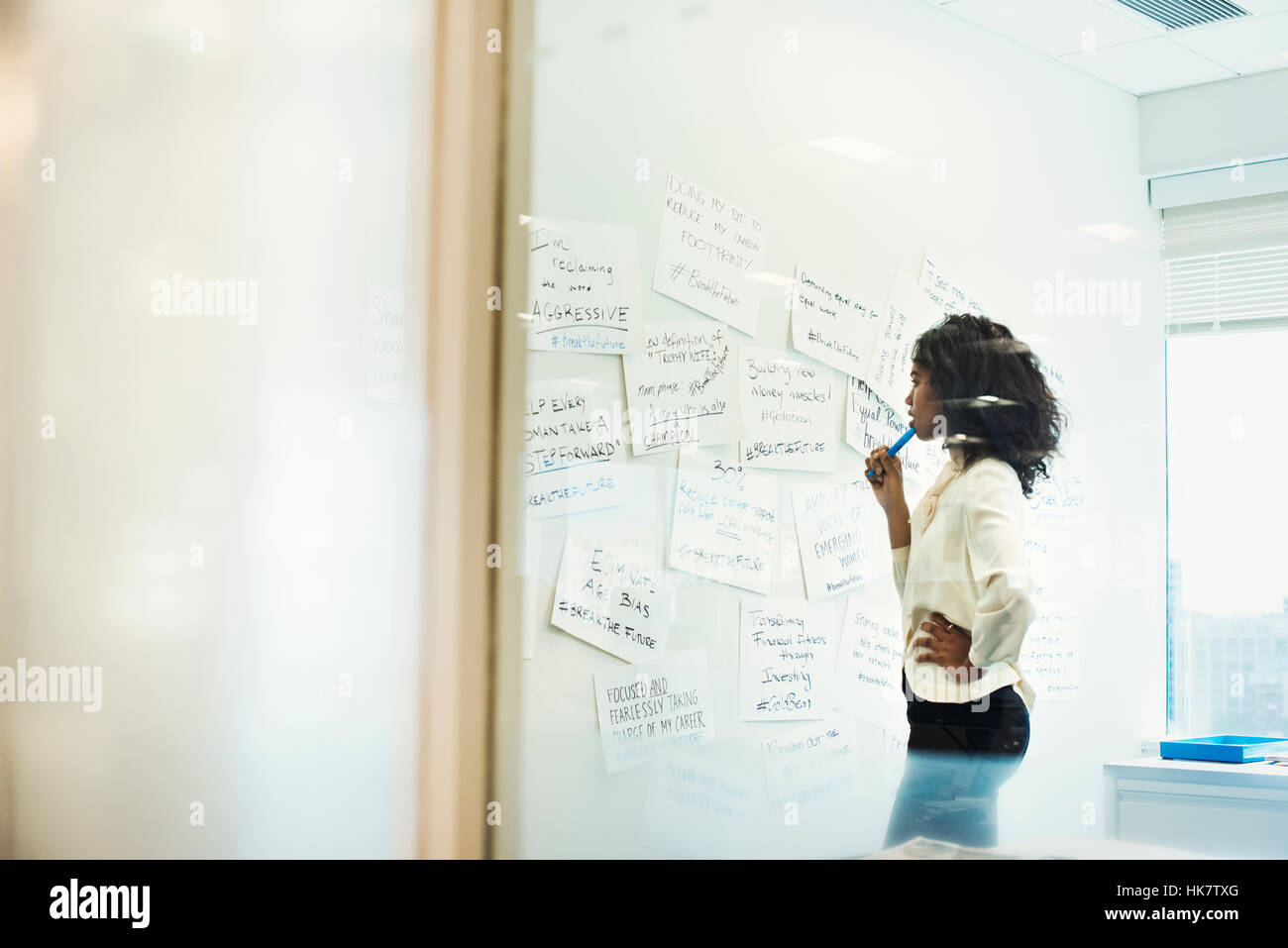 A woman standing in an office looking at pieces of paper pinned on a whiteboard. Stock Photo