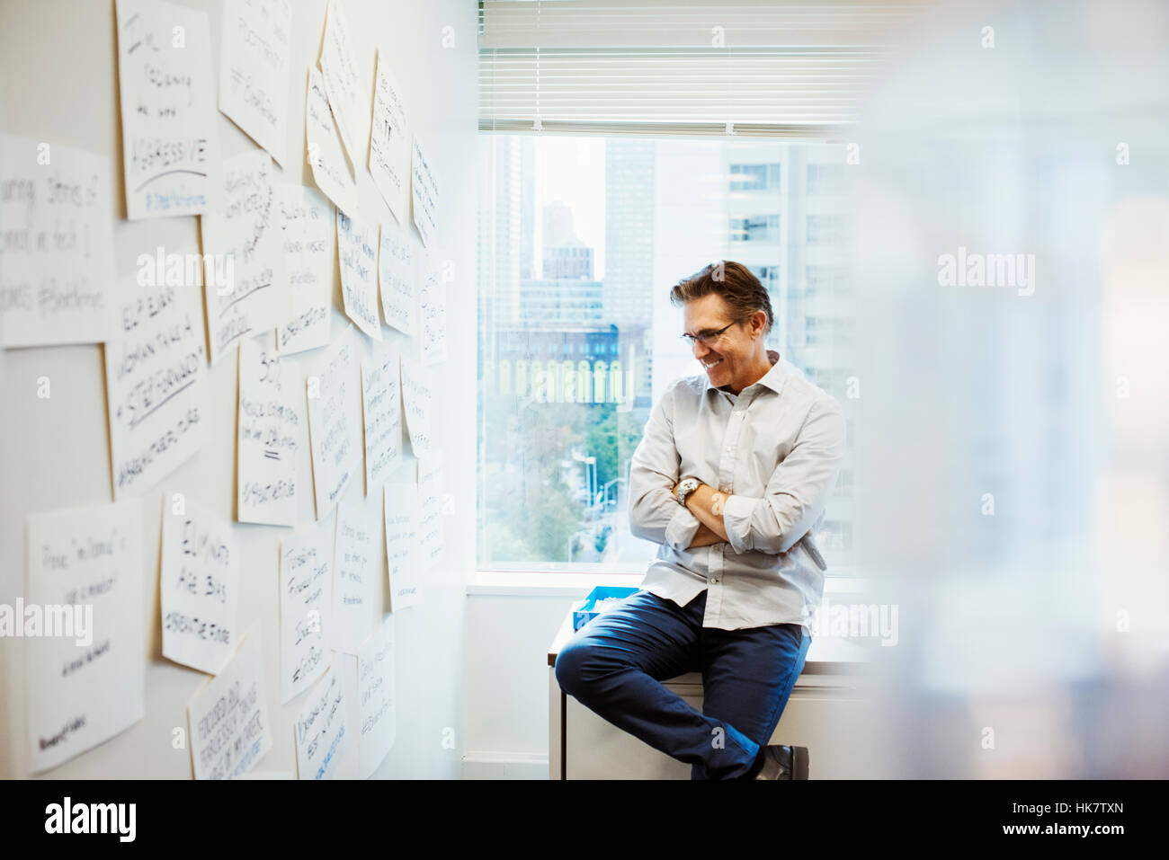 A man leaning on a desk with his arms folded in an office looking at pieces of paper pinned on a whiteboard. Stock Photo