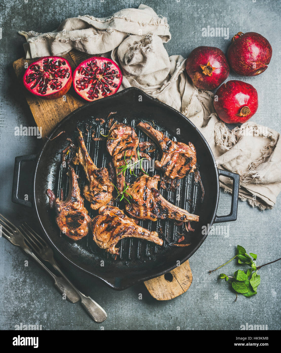Barbecue dinner. Grilled lamb meat chops with onion and rosemary in black cast iron pan served with fresh pomegranates on wooden board over grey concr Stock Photo