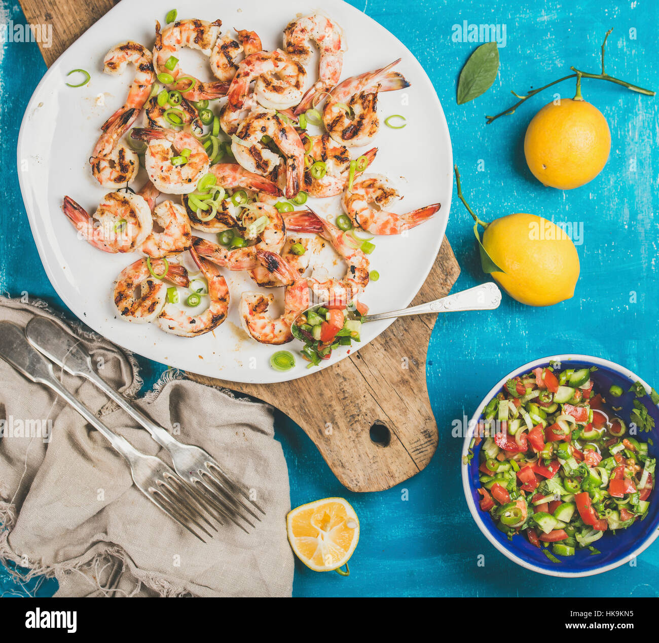 Seafood dinner. Grilled tiger prawns in white plate with lemon, leek, chili pepper and mint salsa sauce on wooden board over bright blue background, t Stock Photo
