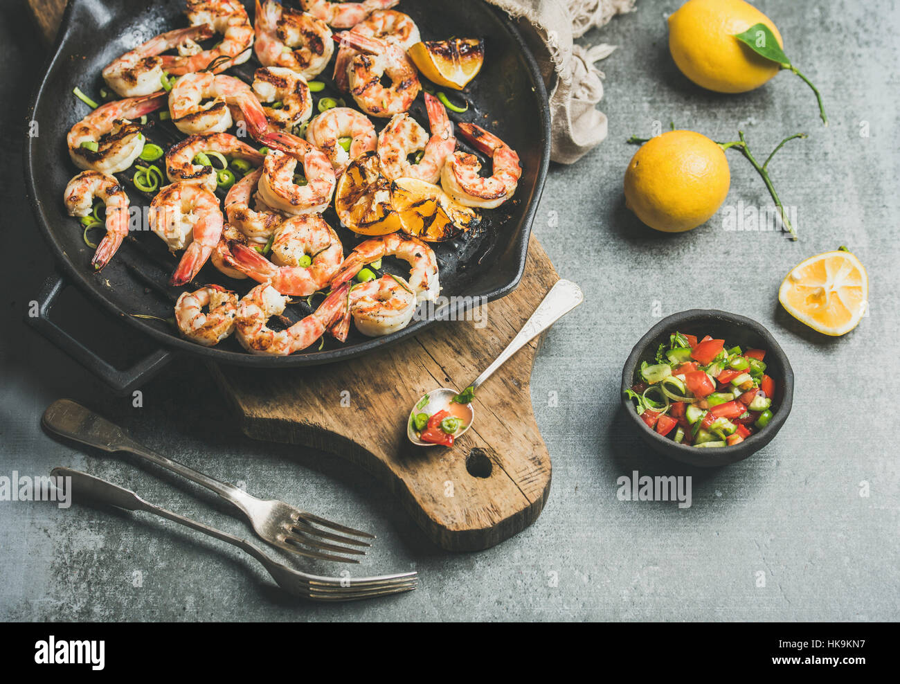 Seafood dinner. Grilled tiger prawns in cast iron grilling pan with lemon, leek, chili pepper and mint salsa sauce over grey concrete background, sele Stock Photo
