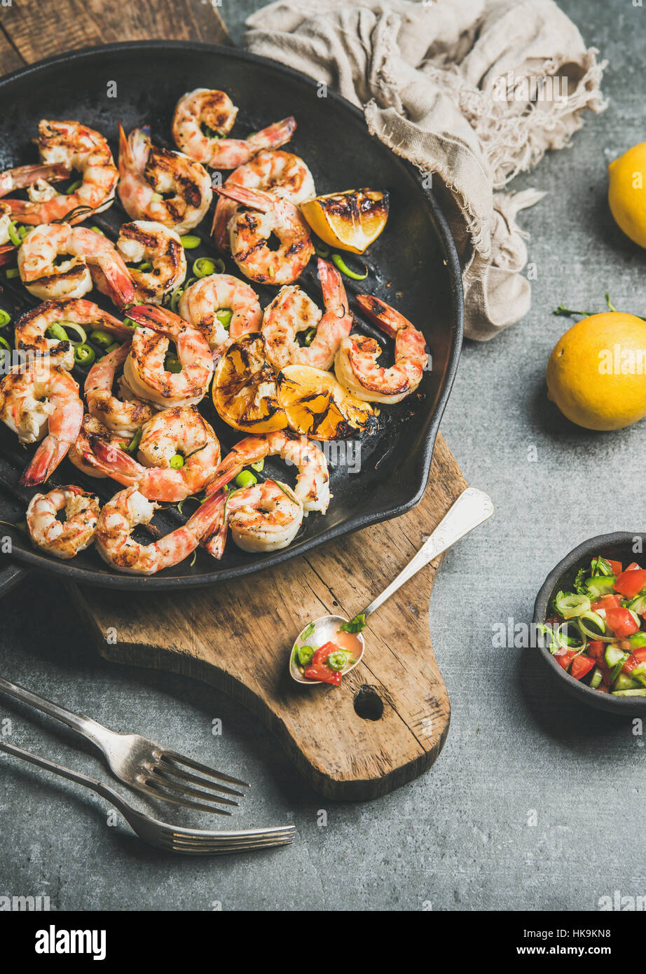 Seafood dinner. Grilled tiger prawns in cast iron grilling pan with fresh lemon, leek, chili pepper and mint salsa sauce over grey concrete background Stock Photo