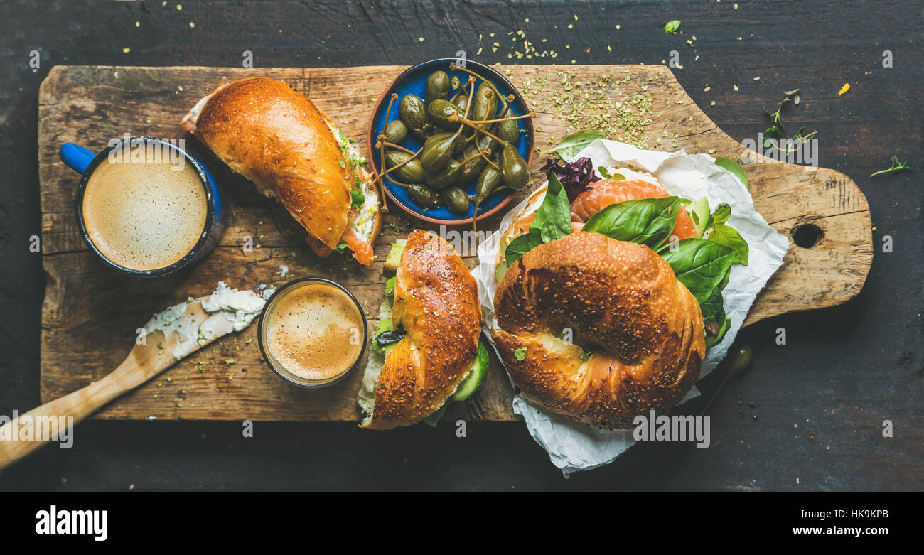 Breakfast with bagel with salmon, avocado, cream-cheese, basil, espresso coffee, capers in blue bowl, rustic wooden board over dark scorched backgroun Stock Photo