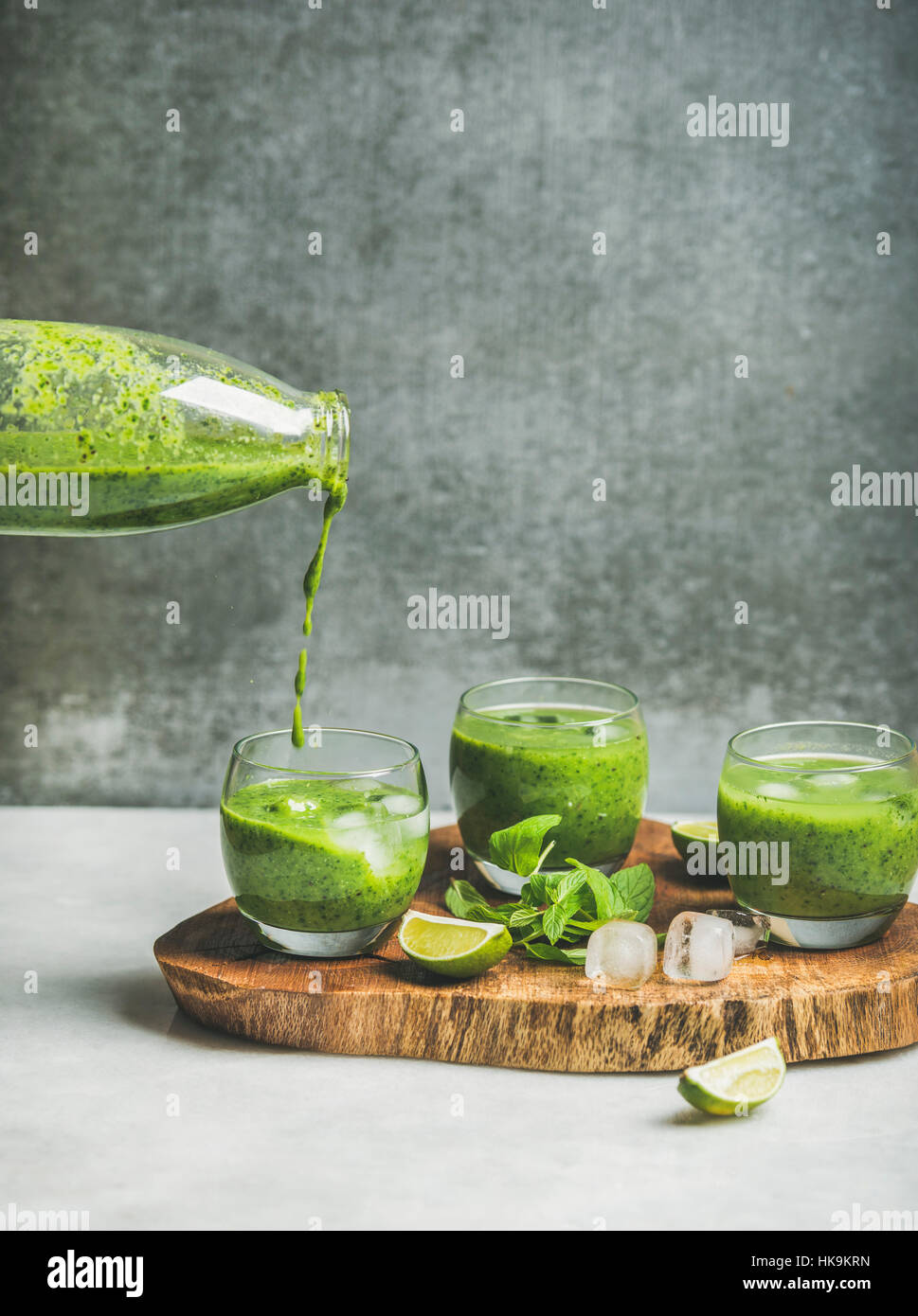 Fresh green smoothie in glasses and pouring from bottle with ice cubes, mint, lime on wooden board, grey concrete wall at background, selective focus, Stock Photo
