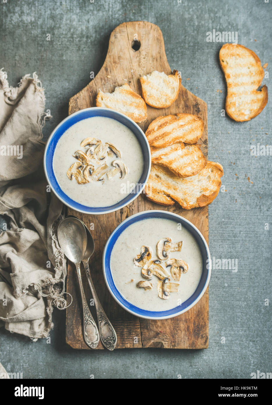 Creamy mushroom soup in bowls with toasted bread slices on rustic serving board over grey concrete background, top view Stock Photo