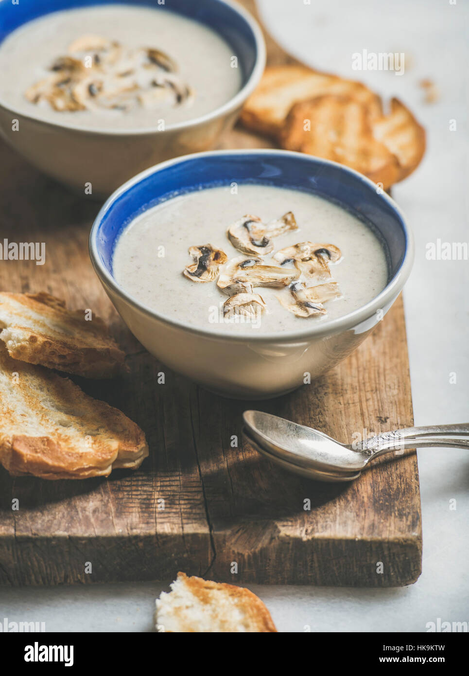 Healthy dinner with creamy mushroom soup in bowls and grilled bread slices on rustic serving board over grey marble background, selective focus Stock Photo