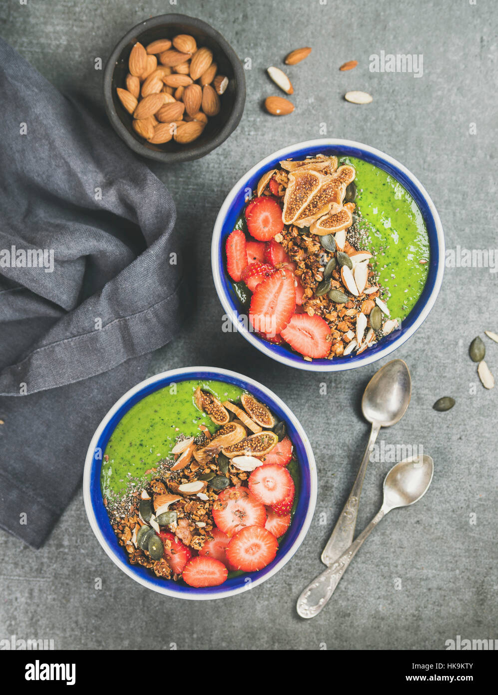 Healthy breakfast. Green smoothie bowls with strawberries, granola, chia and pumpkin seeds, dried figs and almonds over grey concrete background, top Stock Photo