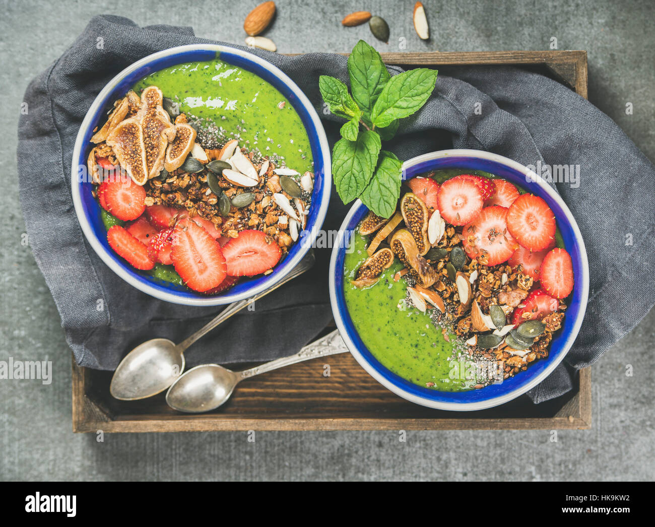 Healthy breakfast. Green smoothie bowls with strawberries, granola, chia and pumpkin seeds, dried figs and nut in wooden tray over grey concrete backg Stock Photo