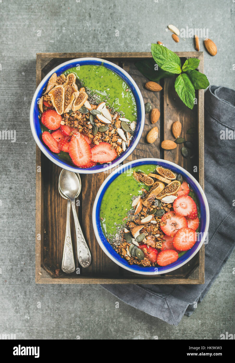 Healthy breakfast. Green smoothie bowls with strawberries, granola, chia and pumpkin seeds, dried fruit and nut in wooden tray over grey concrete back Stock Photo