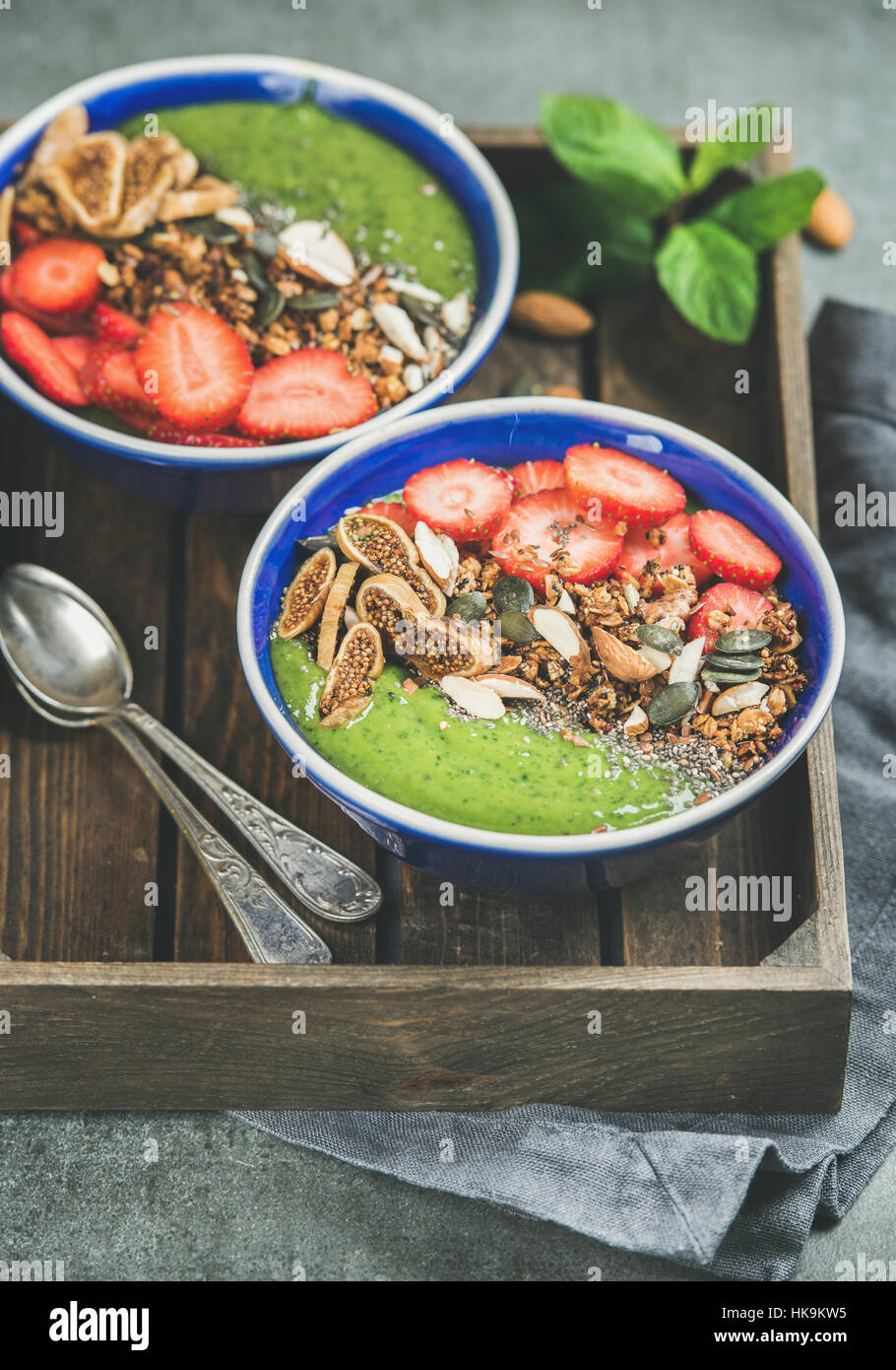 Healthy breakfast. Green smoothie bowls with strawberries, granola, chia and pumpkin seeds, dried fruit and nut in wooden tray over grey concrete back Stock Photo