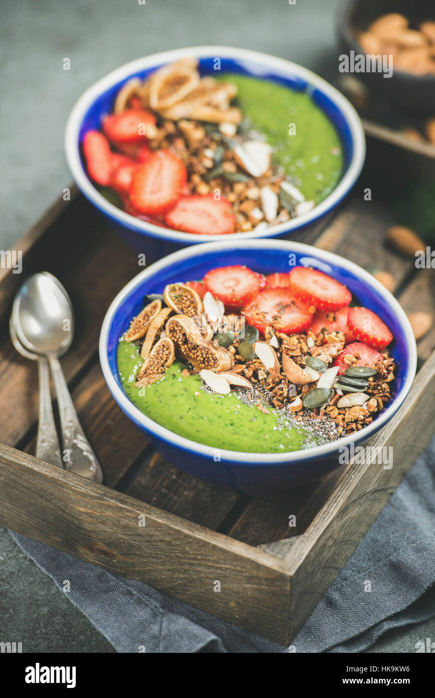 Healthy breakfast. Green smoothie bowls with fresh strawberries, granola, chia and pumpkin seeds, dried fruit and nuts in wooden tray over grey concre Stock Photo