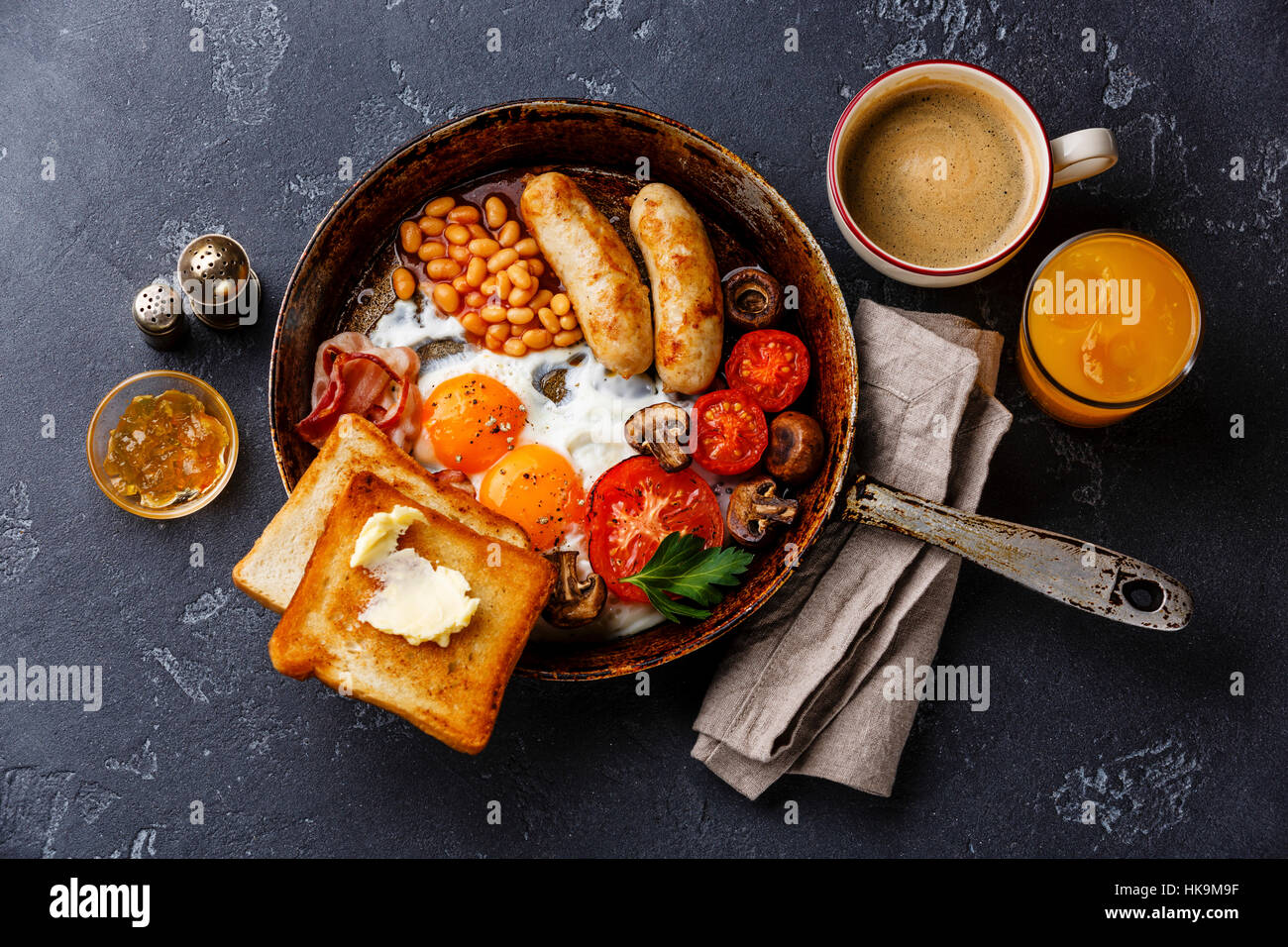 English breakfast in pan with fried eggs, sausages, bacon, beans, toasts and coffee on dark stone background Stock Photo