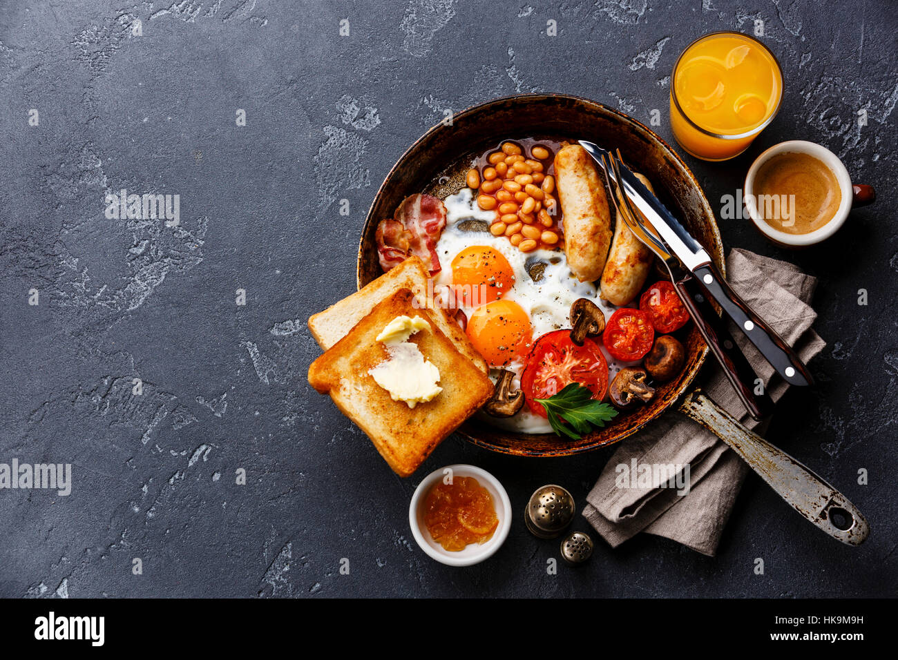 English breakfast in pan with fried eggs, sausages, bacon, beans, toasts and coffee on dark stone background copy space Stock Photo