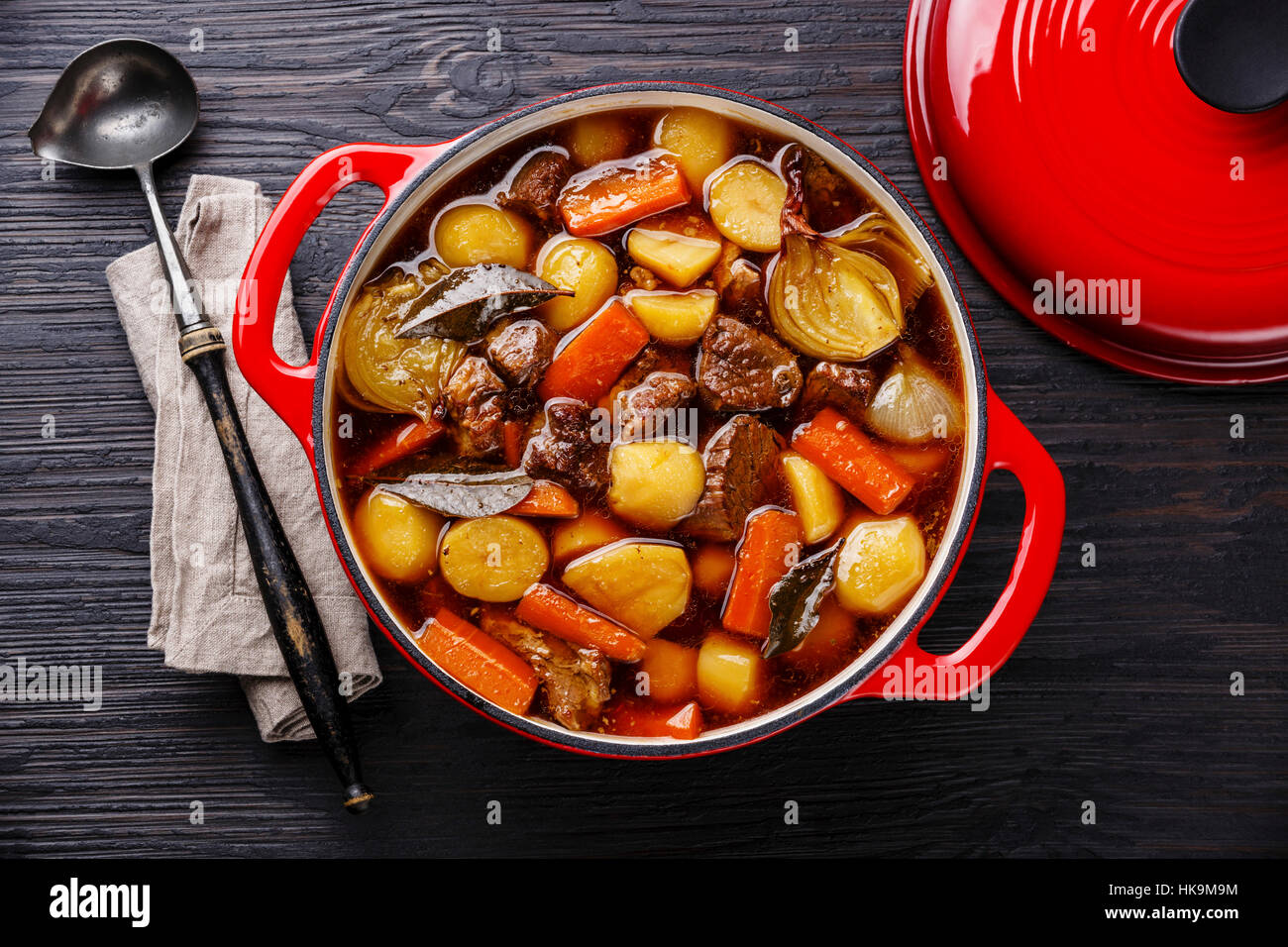 Beef meat stewed with potatoes, carrots and spices in cast iron pot on burned black wooden background Stock Photo